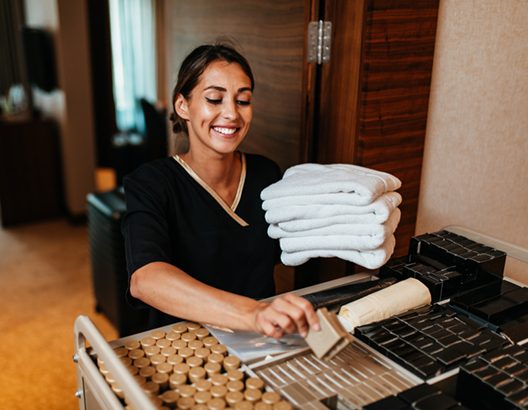 Beautiful young hotel chambermaid in uniform bringing clean towels and other supplies to hotel room.